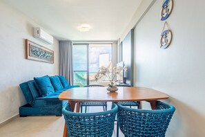 A room with a blue sofa, a wooden dining table, wicker chairs, a framed artwork, and a ceiling light. - Apê Completo em Porto de Galinhas-SH164D (Ipojuca)