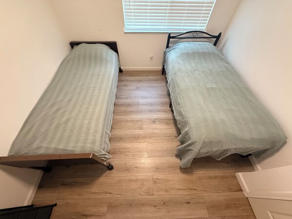 Une petite chambre avec deux lits simples, un plancher en bois et une fenêtre avec des stores.