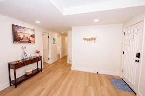 A hallway with wooden flooring, a console table with decorative items, a painting on the wall, and a door with a patterned design. - Family friendly 3-bedroom house with AC in Huntington Beach (Huntington Beach)