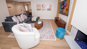 Ett modernt vardagsrum med en grå soffa, en vit fåtölj och ett soffbord. På väggen hänger tre inramade konstverk och ovanför soffan finns en stor tavla med blommotiv.