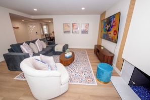 Ein modernes Wohnzimmer mit einem grauen Sofa, einem weißen Sessel und einem Couchtisch. An der Wand hängen drei gerahmte Kunstwerke, darüber am Sofa eine große Blumenmalerei.