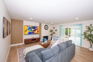 A modern living room with a grey sofa, a wooden TV stand, a colorful abstract painting, and a large window with a view of greenery. - Family friendly 3-bedroom house with AC in Huntington Beach (Huntington Beach)