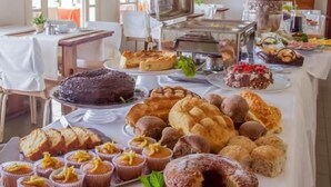 Una mesa de buffet con diversos productos horneados y postres, un televisor y una planta en maceta en una habitación con ventanas amplias y un ventilador de techo.