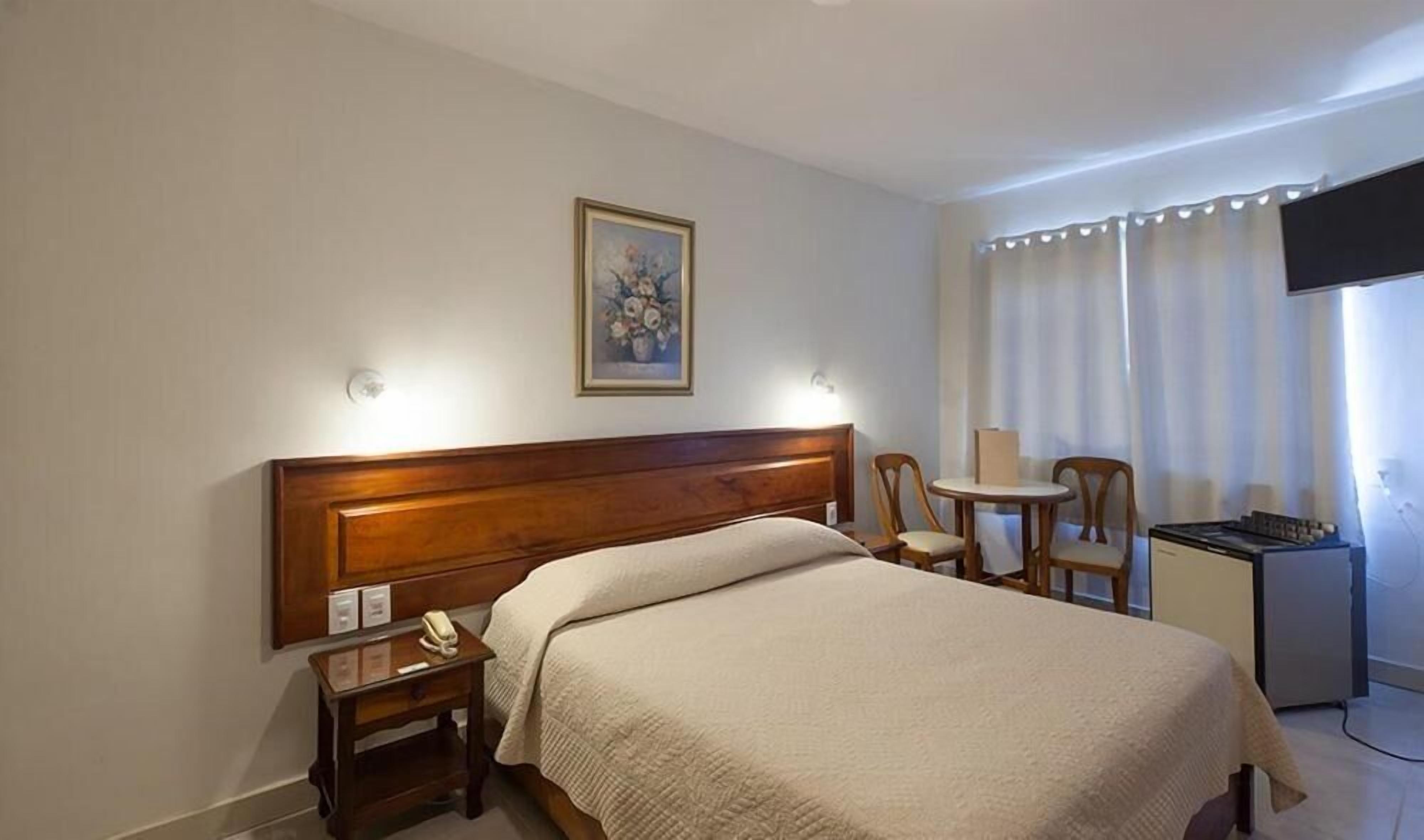 A hotel room with a bed, bedside table, lamp, chair, and a small desk with a television.
