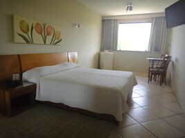 A hotel room with a bed, a desk, a chair, a radiator, and a window with curtains.