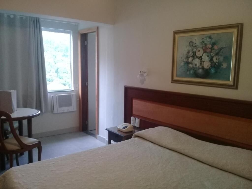 Une chambre d’hôtel avec un lit, une table de chevet, une chaise, une fenêtre avec des rideaux et un tableau au mur.
