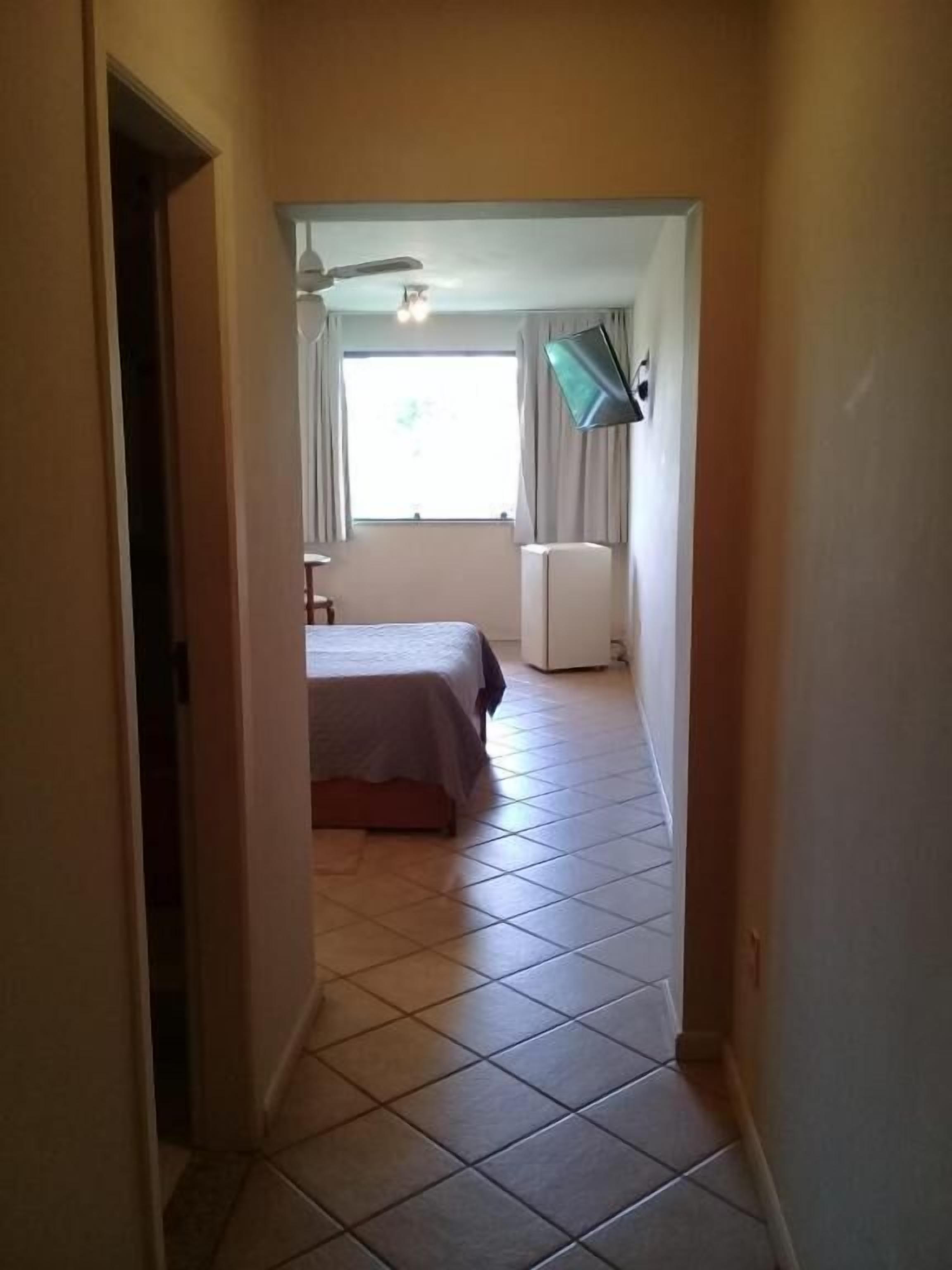 Un couloir étroit mène à une chambre à coucher équipée d’un lit, d’un ventilateur de plafond et d’une fenêtre avec des rideaux.
