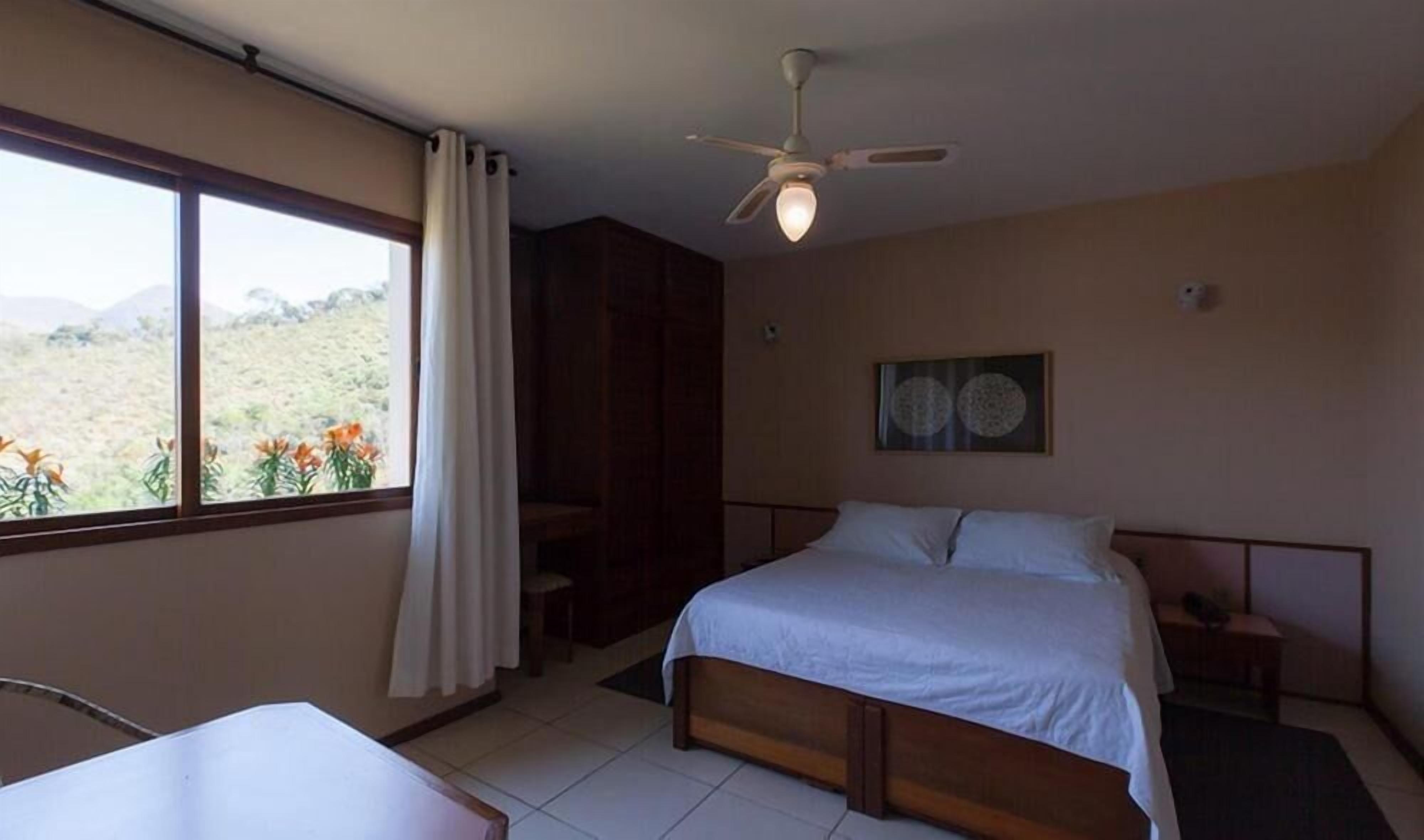 Une chambre à coucher comprenant un lit, un bureau, une chaise, un ventilateur de plafond et une fenêtre avec des rideaux.