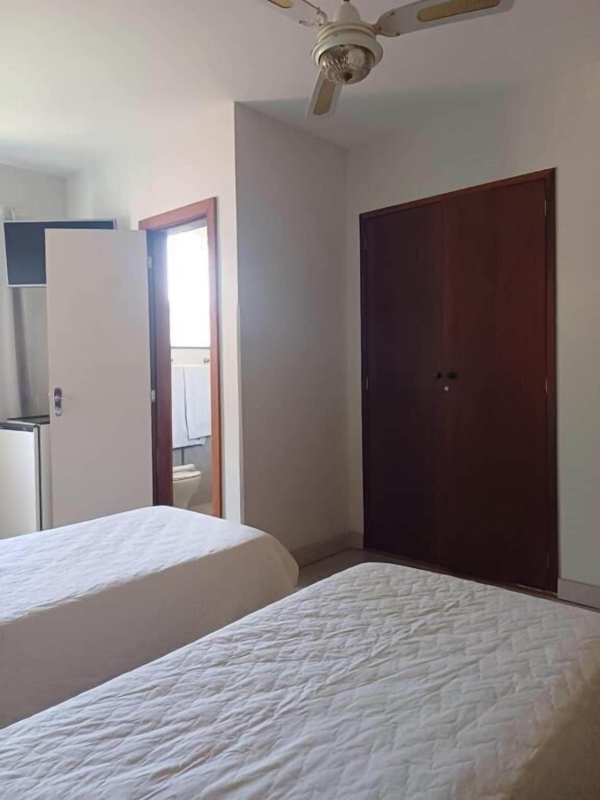 Habitación con dos camas, ventilador de techo, puerta de madera y baño a la vista.