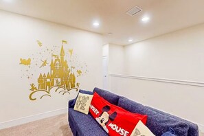 A room with a white wall featuring a castle and mouse decoration, a dark blue sofa with Mickey Mouse pillows, and a white door in the background. - Family Pool Home Near Disney Parks & Golf Fun (Clermont)