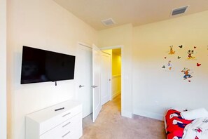 A bedroom with a TV mounted on the wall, a white dresser, a bed with Mickey Mouse-themed bedding, and a hallway leading to another room. - Family Pool Home Near Disney Parks & Golf Fun (Clermont)