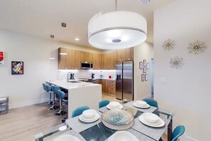 A modern kitchen with a central island, stainless steel appliances, and a dining area with a glass table and blue chairs. - Private Pool & Patio  3BR Family Oasis By Disney (Clermont)