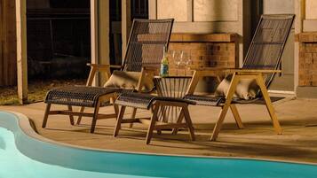 Un espace aménagé au bord de la piscine, avec des chaises longues et une table, une cheminée en briques et une vue dégagée sur la piscine.