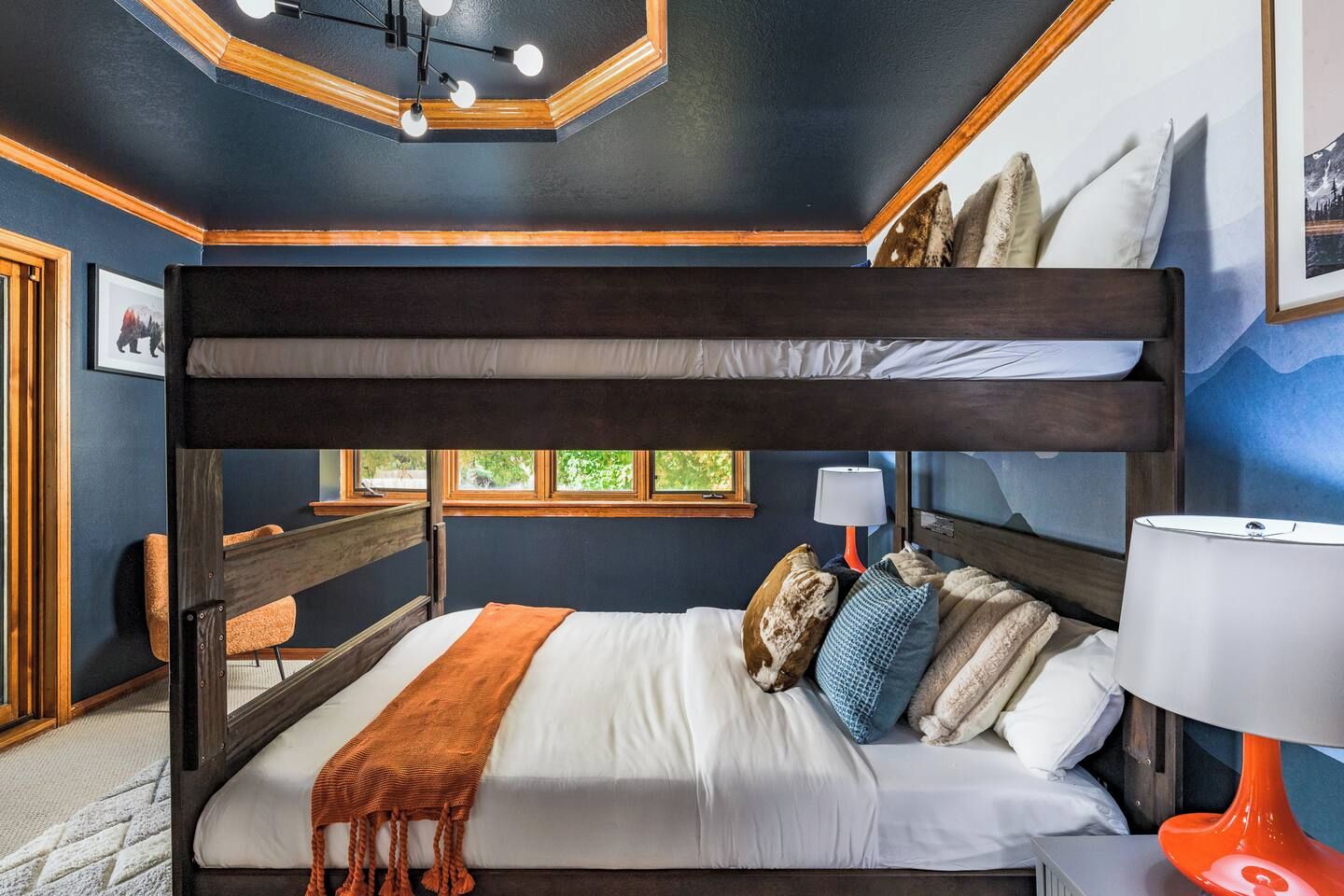 A bunk bed with a wooden headboard, white bedding, and a variety of pillows. There is a desk with a chair and a lamp to the right.