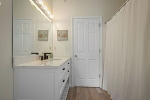 A bathroom with a white vanity, a mirror with lights, a door, and a shower curtain. - Stress-Free Family Beach Stay, Pool + Perks in Rehoboth Beach (Rehoboth Beach)