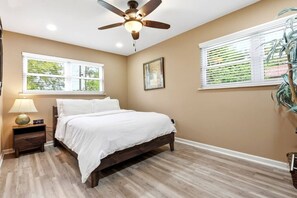 Quarto com cama, mesa de cabeceira, abajur, ventoinha no teto, janelas e uma planta.