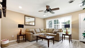 Ein Wohnzimmer mit einer Couch, einem Couchtisch und zwei Beistelltischen, einem Deckenventilator und einem großen Fenster mit Jalousien.