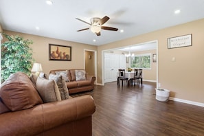 A living room with a brown leather sofa, a ceiling fan, a dining area with a table and chairs, and a hallway leading to a door. - Peaceful North Little Rock Home w Backyard! (North Little Rock)