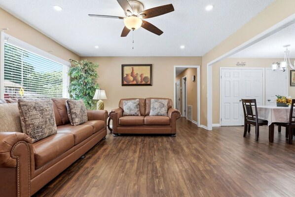 A living room with two brown leather sofas, a ceiling fan, a dining table with chairs, and a hallway leading to a door. - Peaceful North Little Rock Home w Backyard! (North Little Rock)
