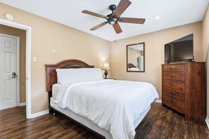 Een slaapkamer met een bed, een houten dressoir, een plafondventilator en een spiegel aan de muur.