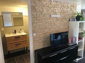 A bathroom with a stone wall, a mirror, a sink with a faucet, and a television on a black stand. - Rosel Apartment, 300 meters from Kersidan Beach (Trégunc)