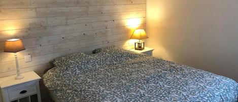 Un dormitorio con cabecera de madera, cama con colcha estampada, dos lámparas de noche y una mesita de noche con cajón.
