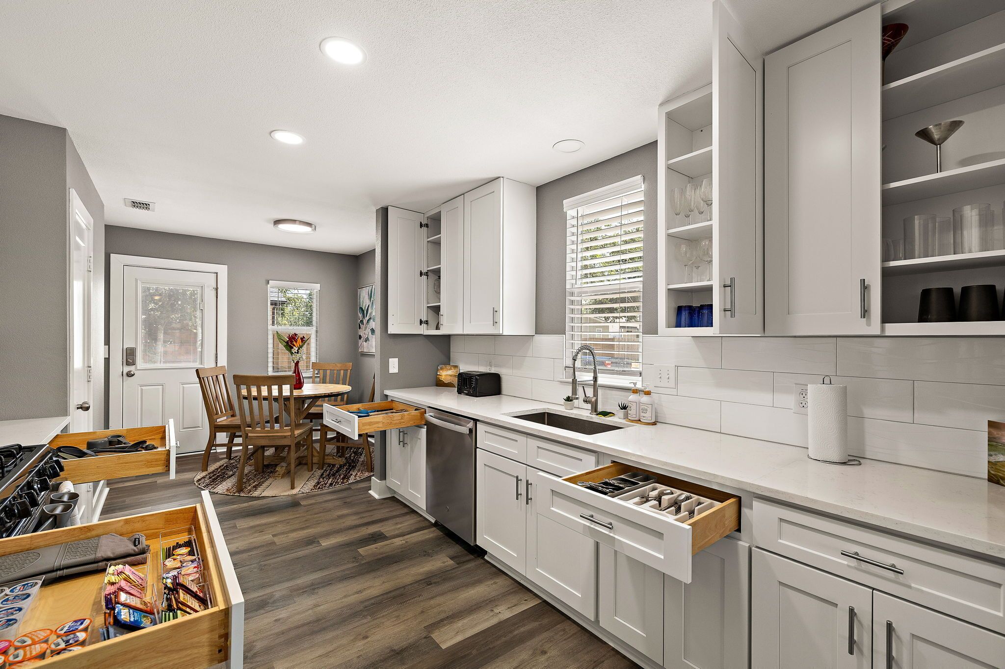 A modern kitchen with white cabinetry, a built-in dishwasher, and a dining area with a wooden table and chairs.