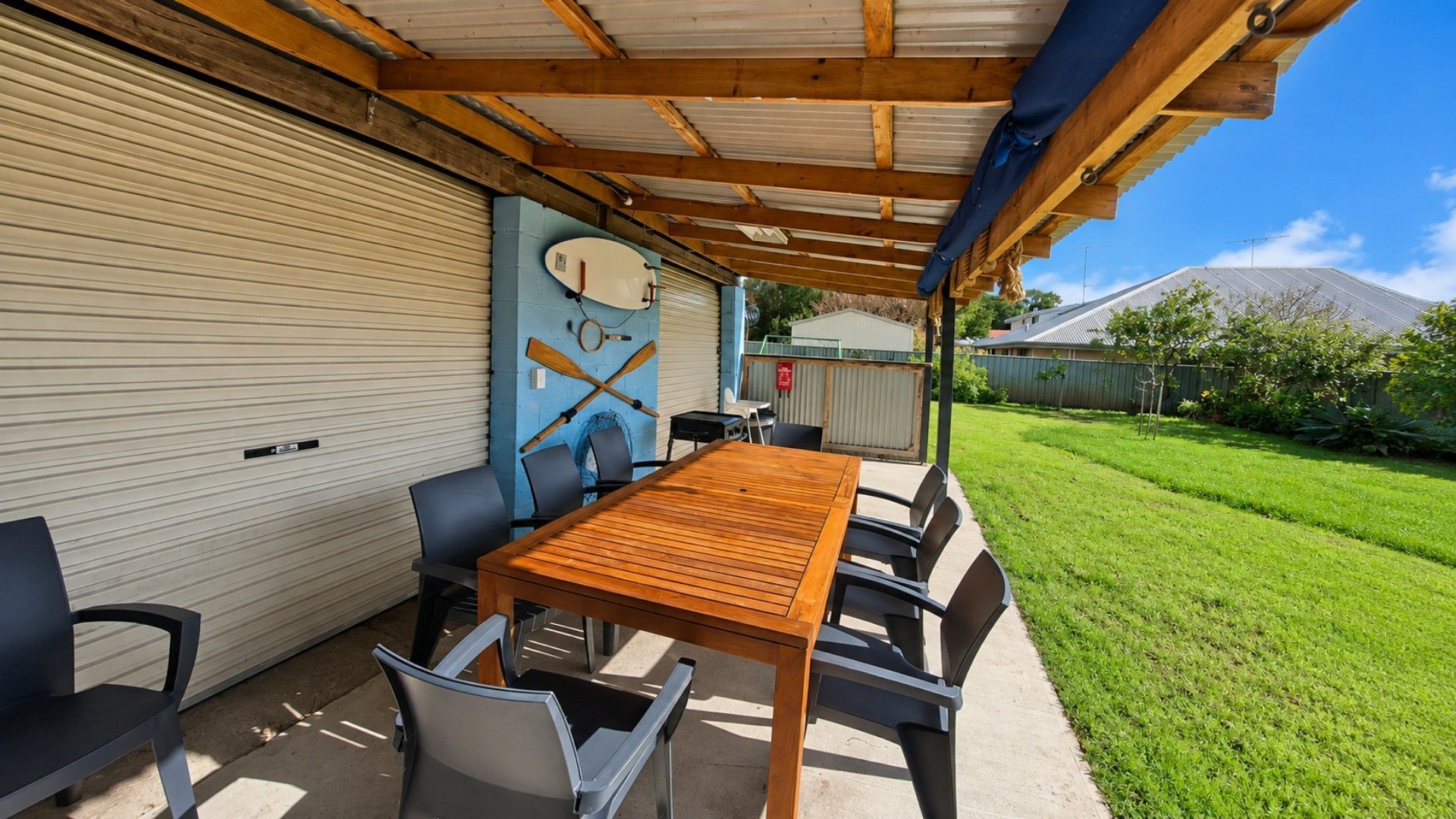 An outdoor patio with a wooden table and chairs, a covered area with a corrugated metal roof, and a garage door.