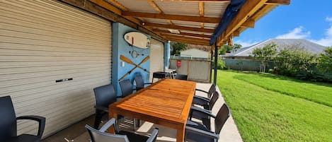 An outdoor patio with a wooden table and chairs, a covered area with a corrugated metal roof, and a garage door.