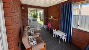 A covered patio area with a brick wall, a sliding glass door leading outside, a white table with chairs, a brown sofa with patterned cushions, and a blue curtain.