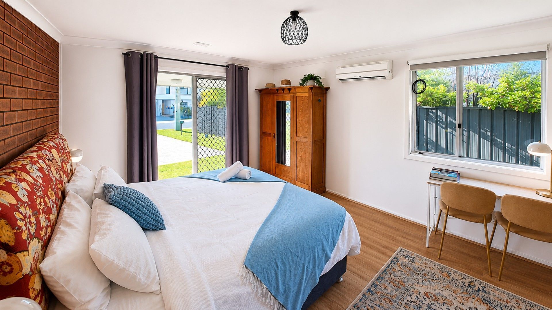 A bedroom with a bed, a desk, chairs, a window, and a view of greenery outside.