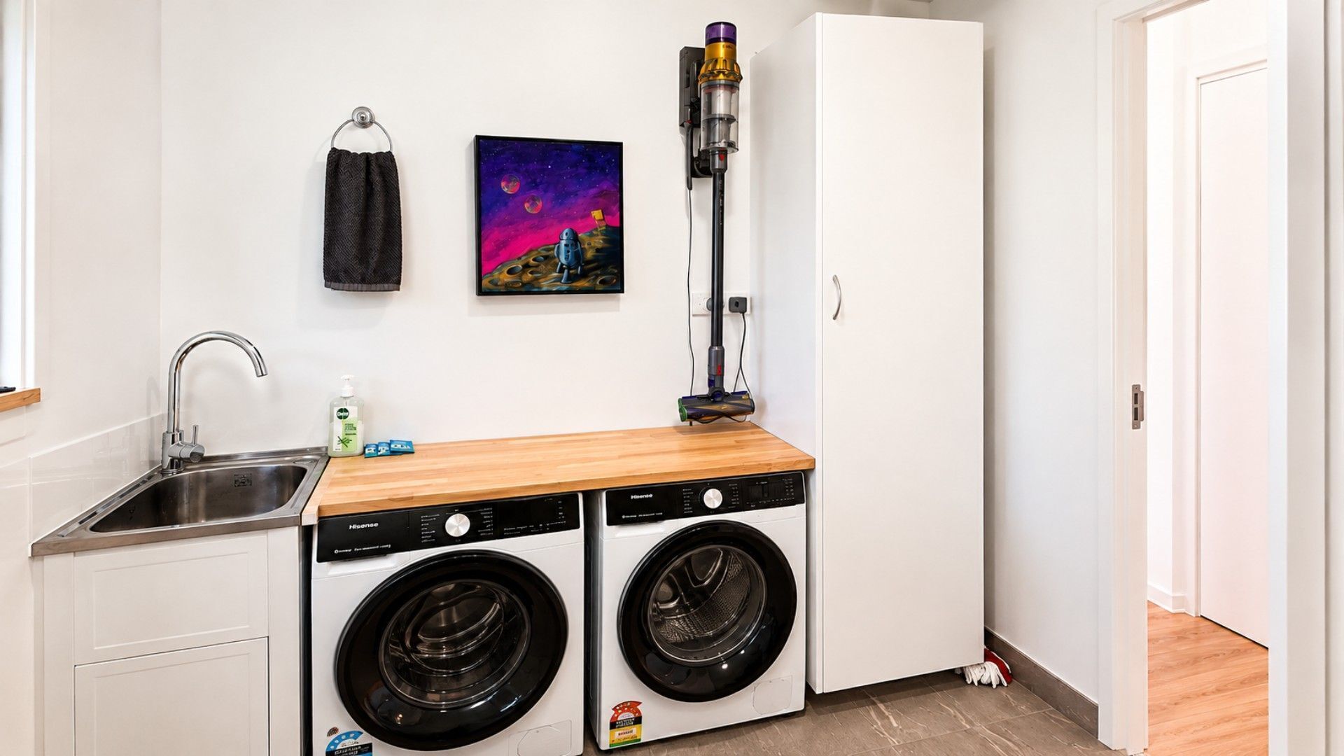 Une petite buanderie comprenant un évier, deux lave-linge à chargement frontal, un plan de travail en bois, un aspirateur et un tableau accroché au mur.