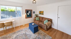 A living room with a sofa, a coffee table, a desk with chairs, a window, and artwork on the walls.