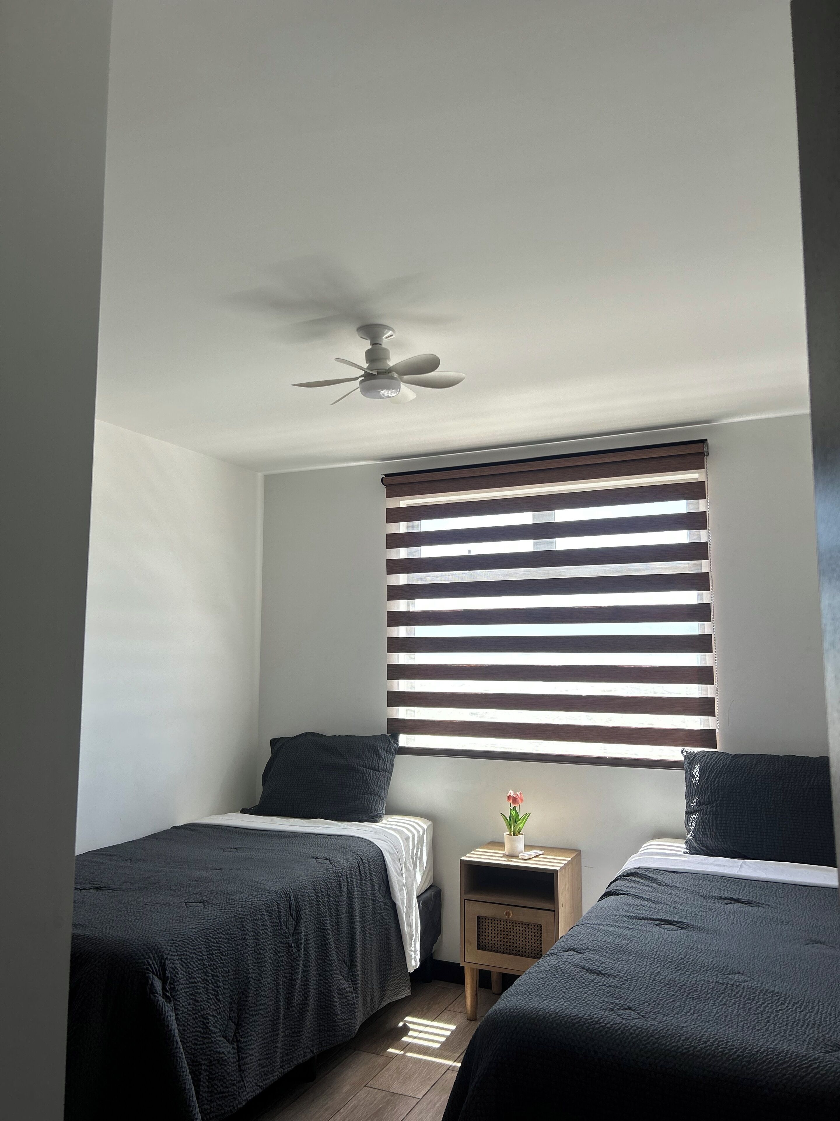 A bedroom with two beds, a ceiling fan, a nightstand with a lamp, and a window with blinds.