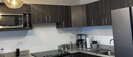 Una cocina moderna con armarios de madera oscura, encimera de granito, refrigerador de acero inoxidable, microondas empotrado, cafetera e iluminación colgante.