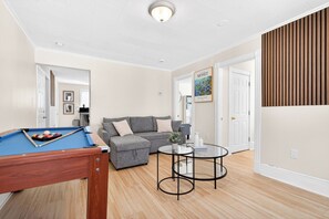 A living room with a pool table, a gray sofa, a glass coffee table, and a door leading to another room.