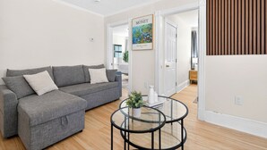 A modern living room with a grey sectional sofa, a glass coffee table, and a framed poster on the wall. - Hidden Pines Retreat - Charming 3 bedroom home near Downtown Boston! (Somerville)