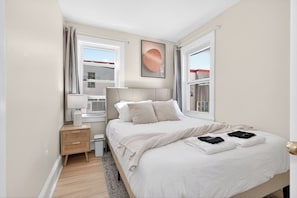 Un dormitorio con cama, mesitas de noche, lámpara, un cuadro en la pared y ventanas.