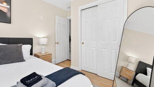 A hotel room with a bed, bedside table, lamp, and a large mirror. - Hidden Pines Retreat - Charming 3 bedroom home near Downtown Boston! (Somerville)