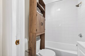 Un baño con un inodoro blanco, un armario de madera, una bañera y paredes cubiertas de azulejos blancos.