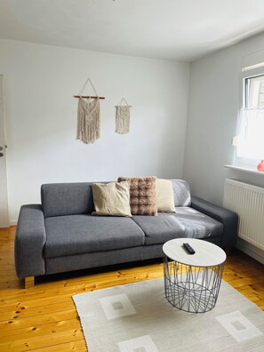 A living room with a grey sofa, two macramé wall hangings, a white round coffee table, and a window with blinds.