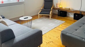 A modern living room with a grey sofa, a wooden floor, a white radiator, a TV mounted on the wall, and a window with curtains. - Lovingly Renovated Apartment in Hausen (Hausen)
