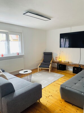 Ein modernes Wohnzimmer mit grauem Sofa, Holzboden, weißem Heizkörper, Wandfernseher und einem mit Vorhängen versehenen Fenster.