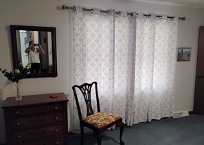 A room with a wooden dresser, a chair with a floral cushion, a mirror, and patterned curtains. - Charming house in tranquil North Western Mesick, Michigan for a relaxing getaway (Mesick)