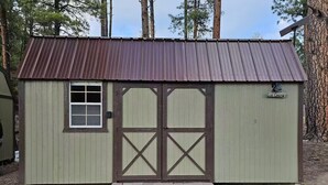 Et lille, etage fritliggende skur med brun dør og vindue, med teksten 'Elk Cabin', beliggende i et skovområde med metal tag og en lille veranda.