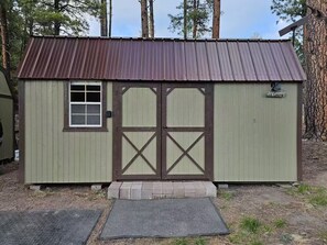 A small, single-story shed with a brown door and window, labeled 'Elk Cabin,' situated in a wooded area with a metal roof and a small porch. - Cozy Alpine Mini Cabin -Elk Cabin- (Alpine)