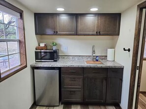 A compact kitchen with dark cabinets, a built-in microwave, a stainless steel dishwasher, and a window providing a view of the outdoors. - Cozy Alpine Mini Cabin -Elk Cabin- (Alpine)