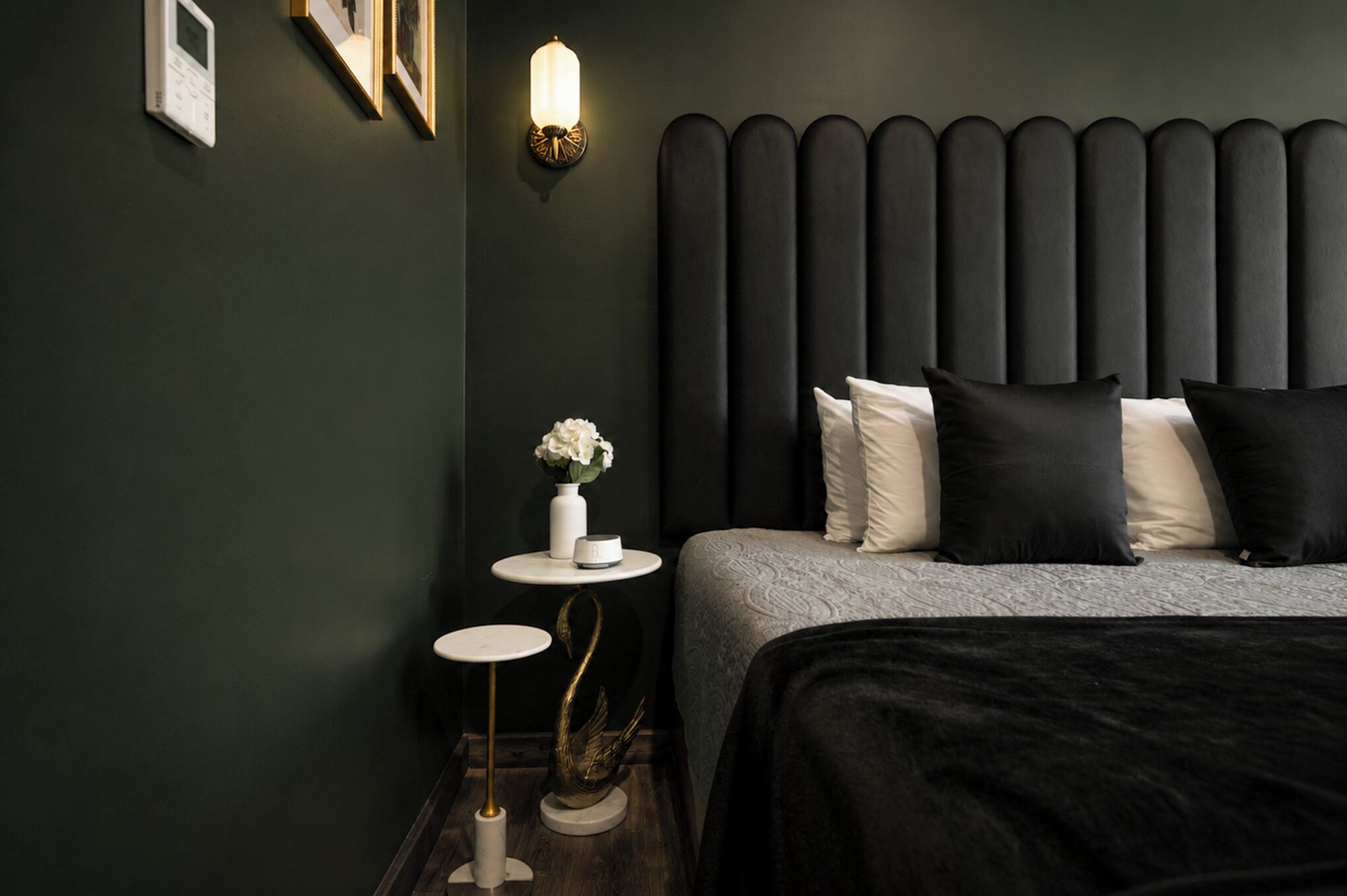 Quarto moderno com parede verde escura, cama com roupa de cama branca e preta, mesa de cabeceira com um vaso de flores e luminária fixada na parede.