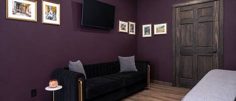 A room with a black sofa, a small table with a glass container, a bed with a patterned blanket, a wooden door, and a mounted TV.