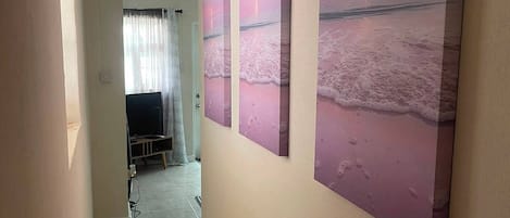 A hallway with a tiled floor, a television in the background, and three beach-themed wall art pieces.